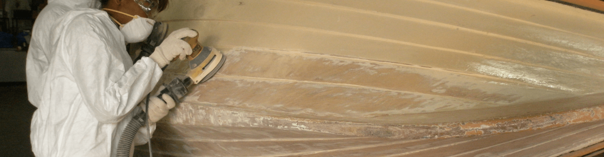 Worker using hull sanding equipment in a boatyard, removing dust during hull repair with flexible and abrasion-resistant tools.