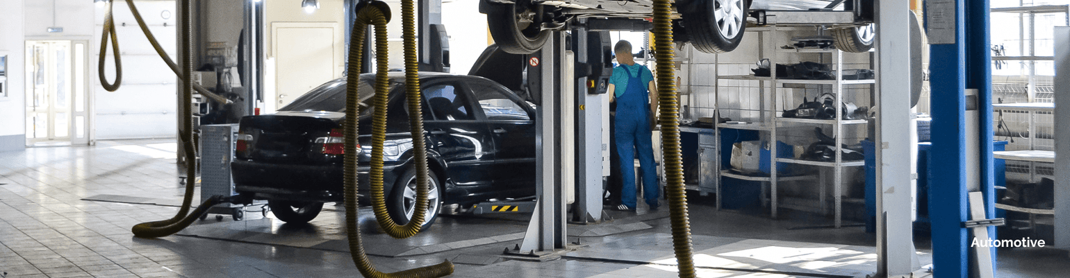 Automotive service bay with a mechanic working on a raised car, exhaust ducts managing fumes in a clean environment.
