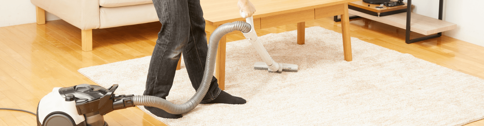 Person vacuuming a carpet with a residential vacuum cleaner in a bright living room setting.