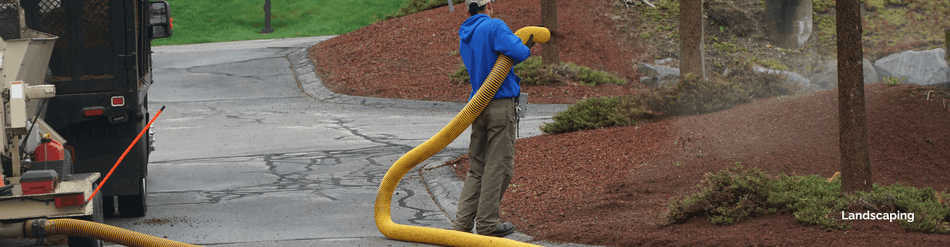 A landscape contractor using a yellow hose for mulch blowing in a well-maintained garden area.